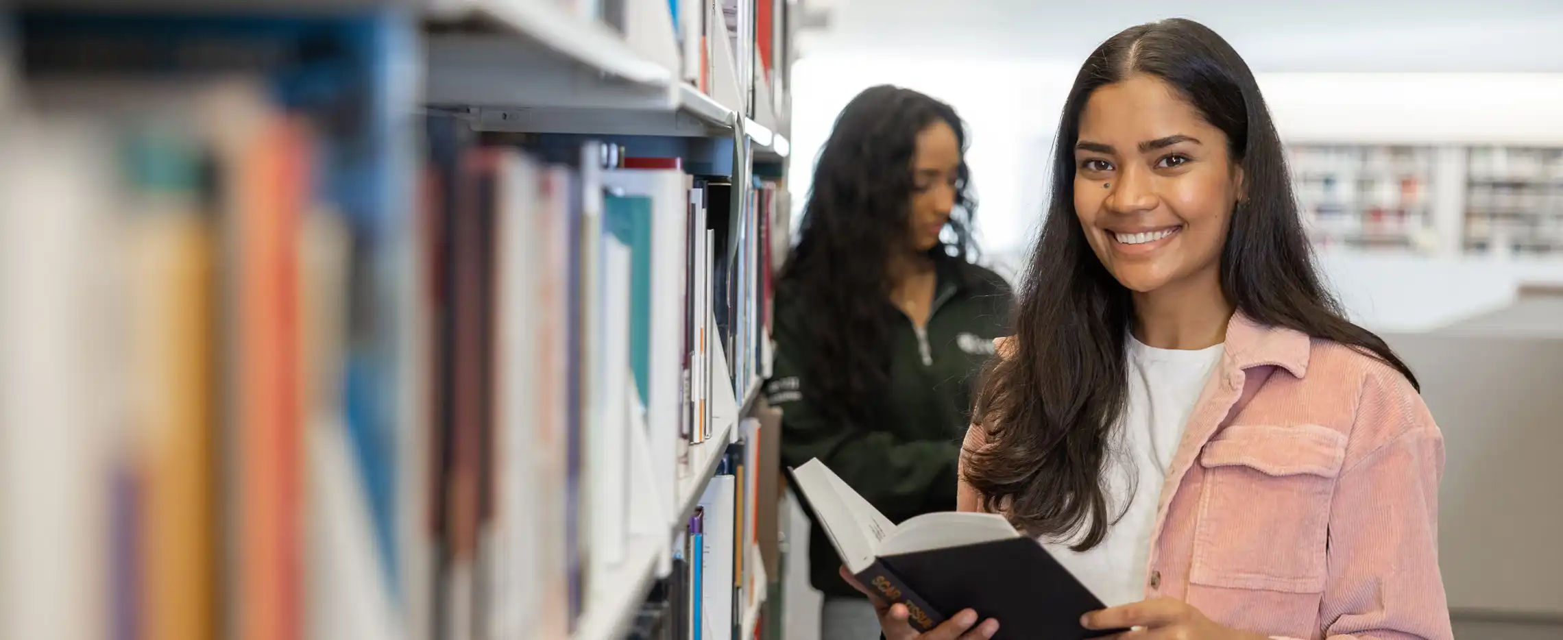 Portrait of smiling university student standing in the library holding a book.