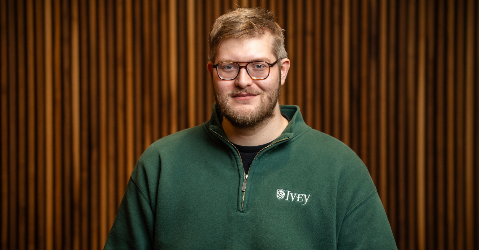 Photo Of Ethan Milne, Ivey Phd Candidate, Wearing Green Ivey Branded Sweater In Wood Paneled Room